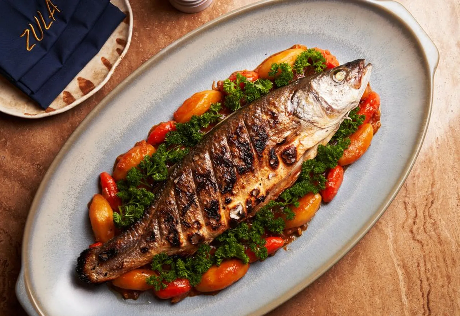 A whole roasted branzino on an oval platter with herbs and tomatoes