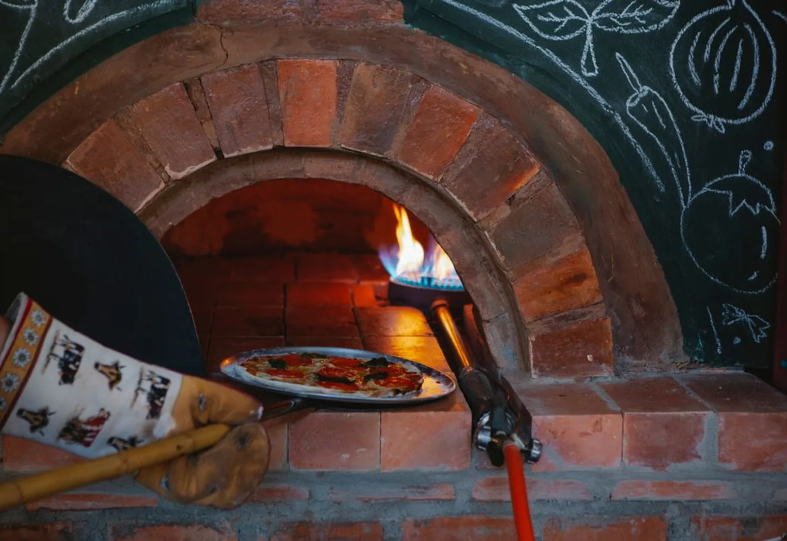 A pizza going into the glowing mouth of a brick wood-fired oven on a long peel